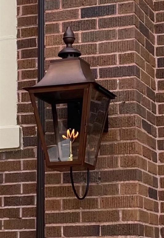 A brown copper outdoor foyer lantern mounted on a brick wall, featuring clear glass and a visible internal flame.