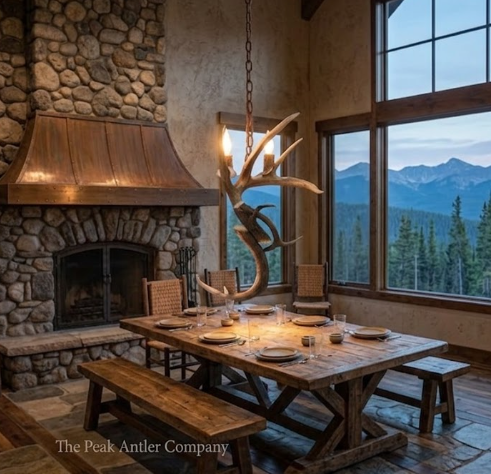 Dining room with stone fireplace, wooden table, and antler chandelier, featuring a mountain view.