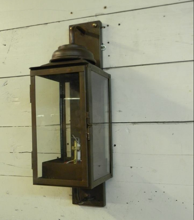 A wall-mounted copper lantern with a clear glass panel, featuring a bronze-like finish, attached to a white wooden surface.