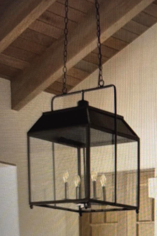 A black metal lantern-style pendant light with a wire mesh screen and candle-style bulbs, hanging from chains in a home interior with exposed wooden ceiling beams.