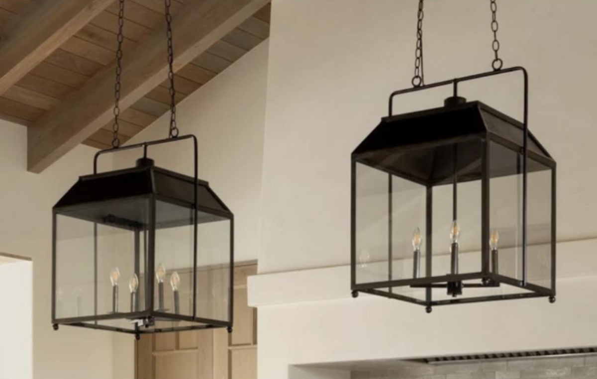 Two black metal lantern-style pendant lights with clear glass panels and visible candle-style bulbs, hanging in a real interior space with exposed wood ceiling beams and cream walls.