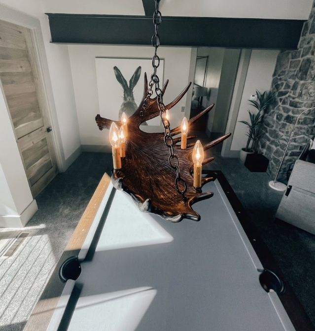 Faux moose antler chandelier hanging above a pool table in a room with stone wall and wooden door.