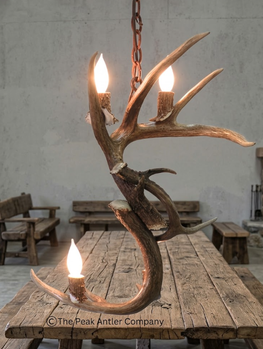 Antler chandelier with three lit bulbs on a rustic wooden table, branded 'The Peak Antler Company'.