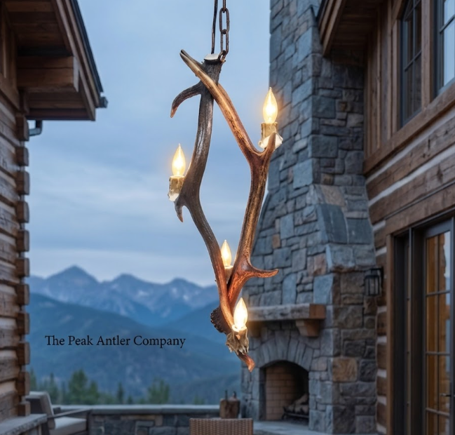 Antler chandelier in front of a stone building with mountains in the background, featuring 'The Peak Antler Company'.