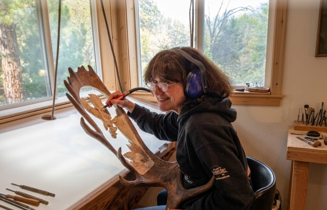 Person working on a wooden sculpture in a workshop with large windows.