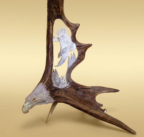 Decorative wall art on a moose antler and eagle sculpture at the base and a soaring eagle in the heart of the antler, on a beige background