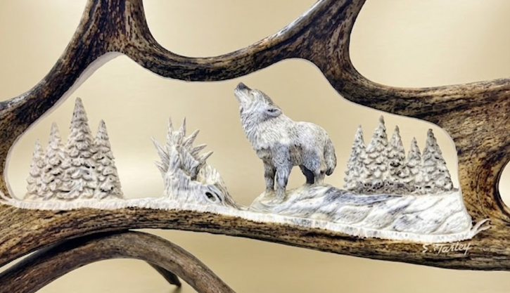 Decorative scene with a wolf howling, trees, and mountains inside moose antlers on a beige background