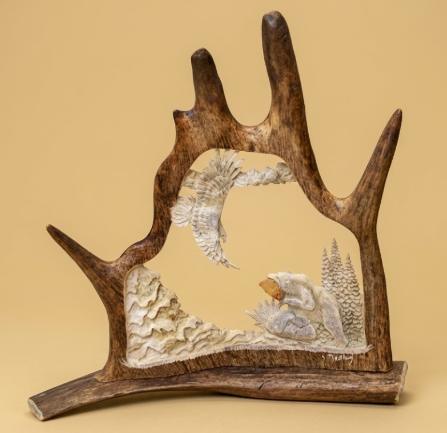 Decorative piece, hand carved into a moose antler, natural elements of a soaring eagle and a hungry fishing bear amongst the forest and river, on a  beige background