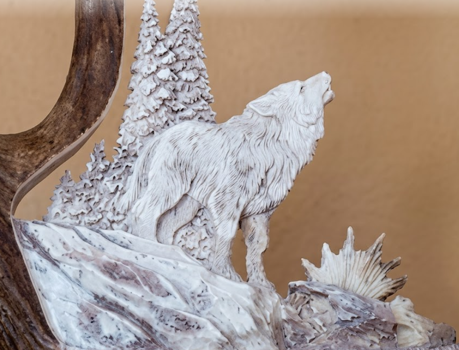 Moose shed carving of a wolf howling at the moon antler carving. Against a tan background.