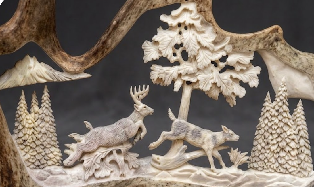 Moose antler carved scene with deer, trees, and mountains on a dark background