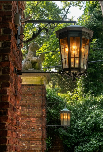 Decorative outdoor black foyer entryway light attached to a brick wall with greenery in the background
