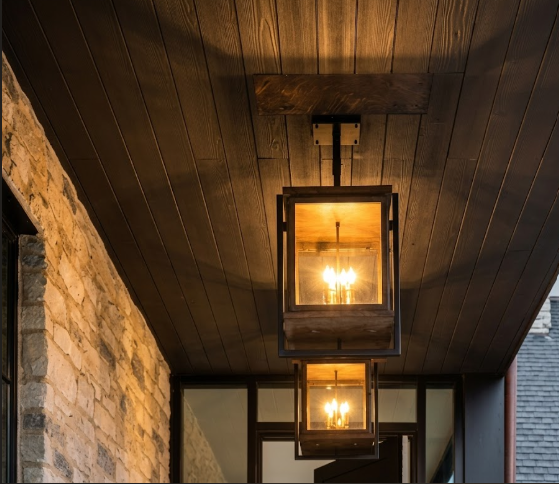 Two hanging decorative foyer entryway pendant lights with glass panels  lights on a wooden ceiling with stone wall and window in the background.